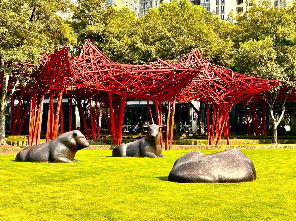 这个场景里的红色装置艺术是 **《城市之林》（也叫 “红亭”）**，由中国艺术家展望创作，以红色钢结构交织成树林形态，是上海静安雕塑公园的标志性装置。
前景的青铜牛雕塑是西班牙艺术家胡安・苏比拉克的作品 **《牛》（或《卧牛》）**，以简洁厚重的造型展现力量感；右侧的石头状雕塑则是同系列的《石》，共同构成充满自然张力的景观。